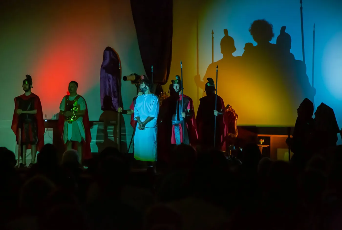 The trial of Christ — a figure in white under blue light, the court's giant shadow thrown across the wall in red, gold and blue
