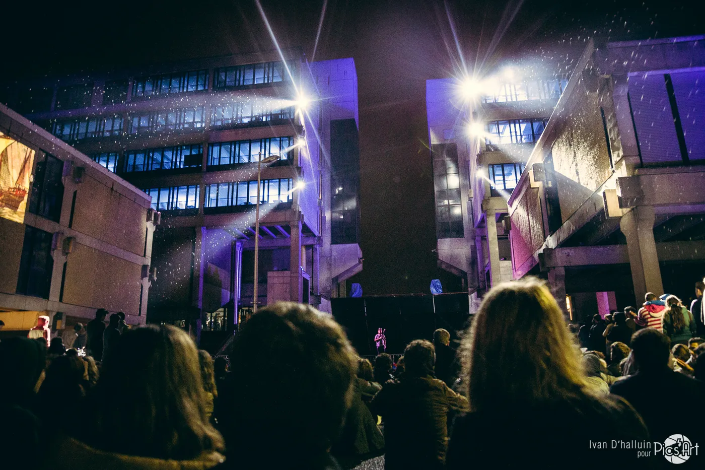 Silhouetted spectators watching buildings washed in violet light, spotlights flaring