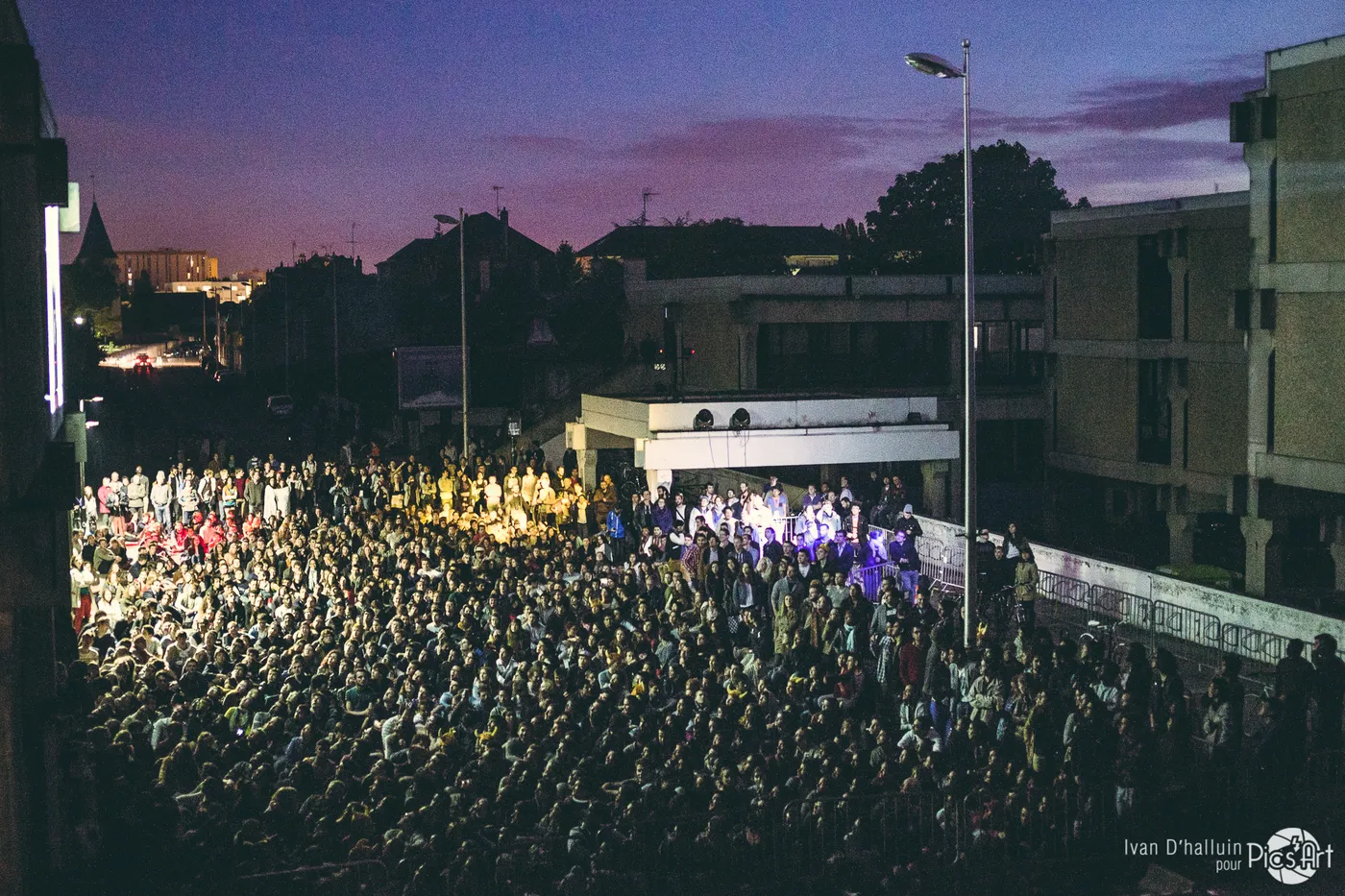 A large seated crowd at dusk before a distant lit stage